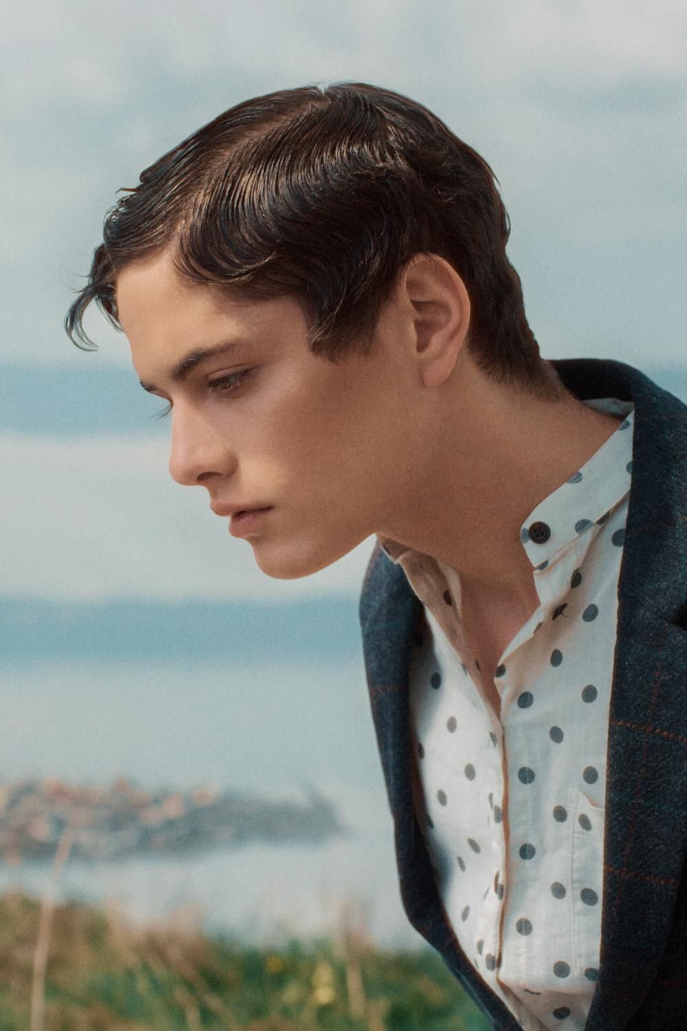 Young man in polka dot shirt and navy blazer gazes down with coastal landscape behind him.