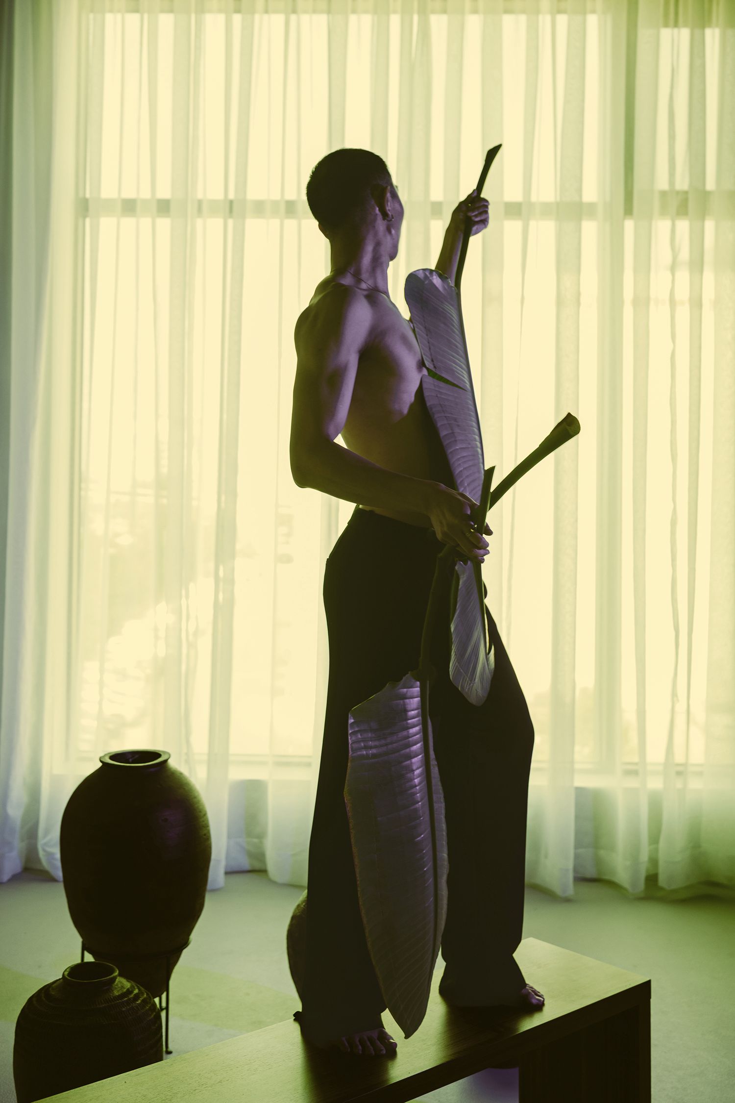 Shirtless man holding traditional weapon stands in silhouette before sheer curtained window.