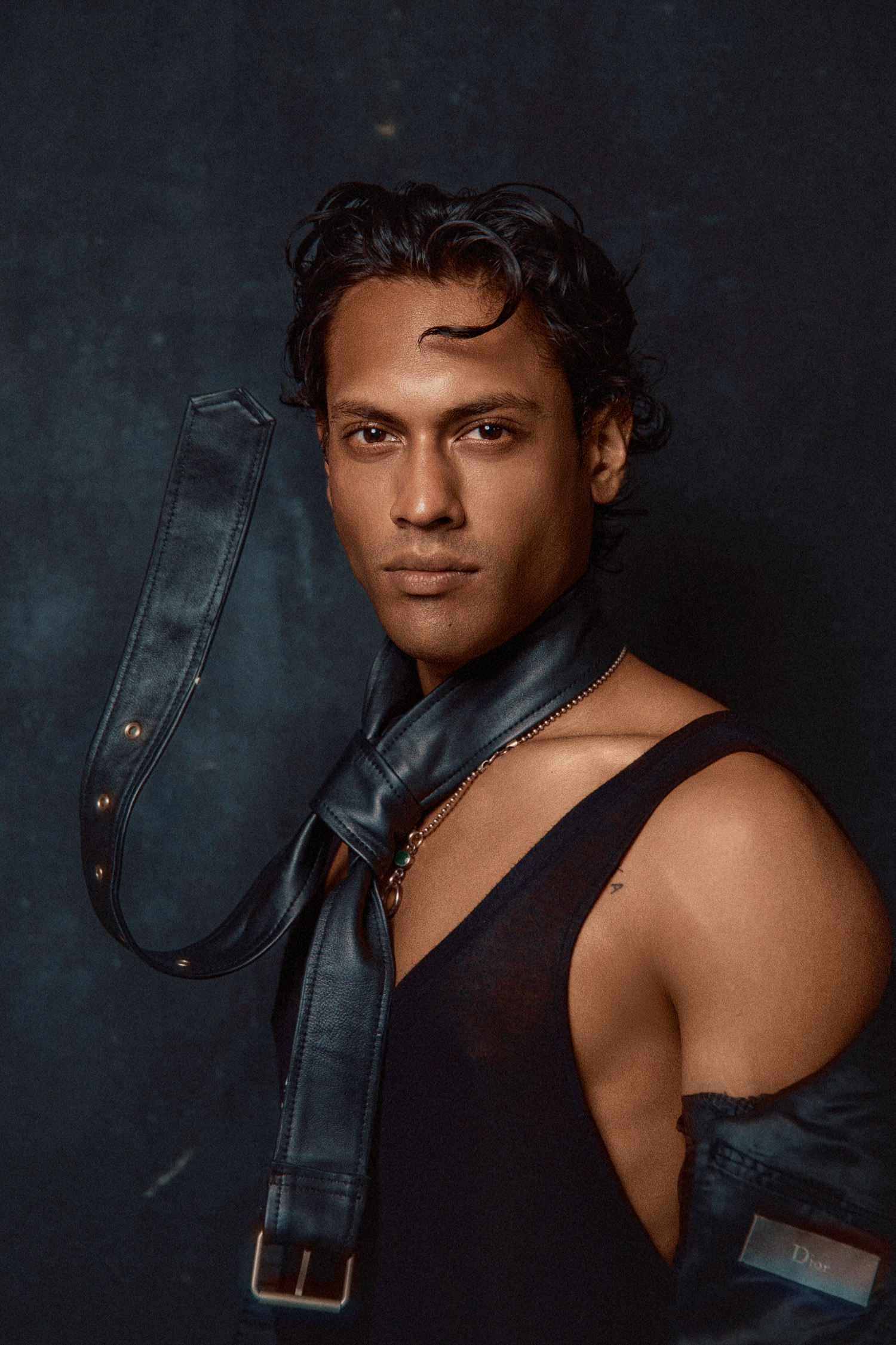 Athletic man in black tank top with leather strap accessory posing against dark textured backdrop.