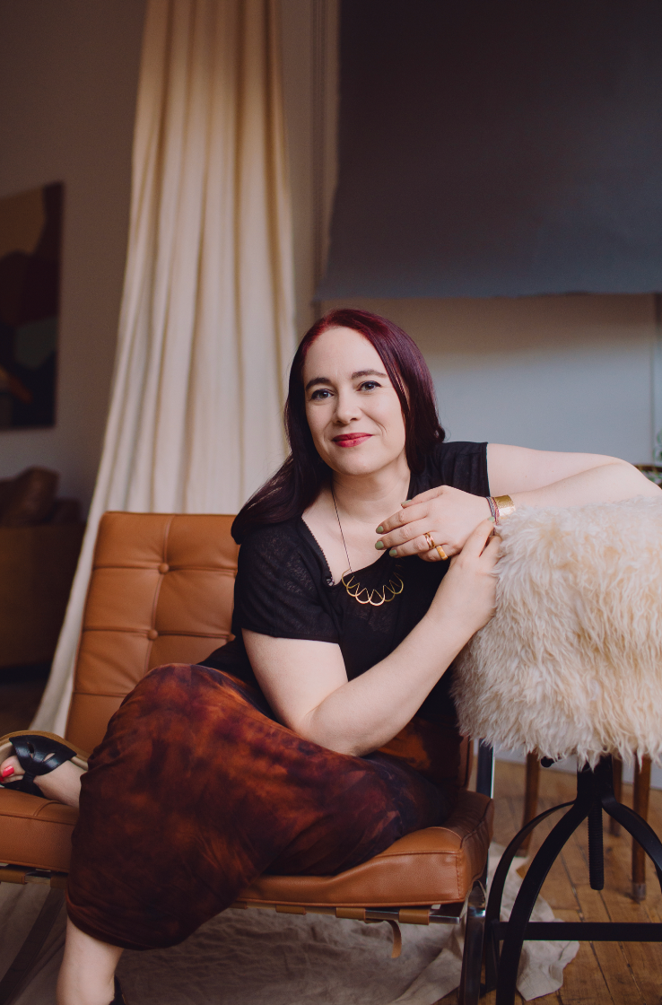Woman with purple hair in black top and burgundy skirt sits relaxed on leather chair in studio.