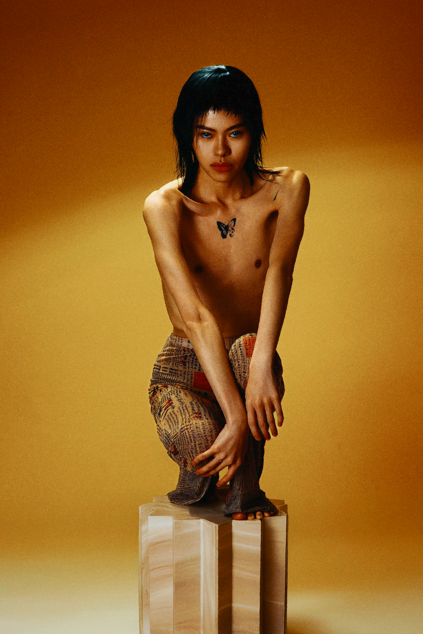 Shirtless model with butterfly tattoo crouching on clear acrylic pedestal against golden background.