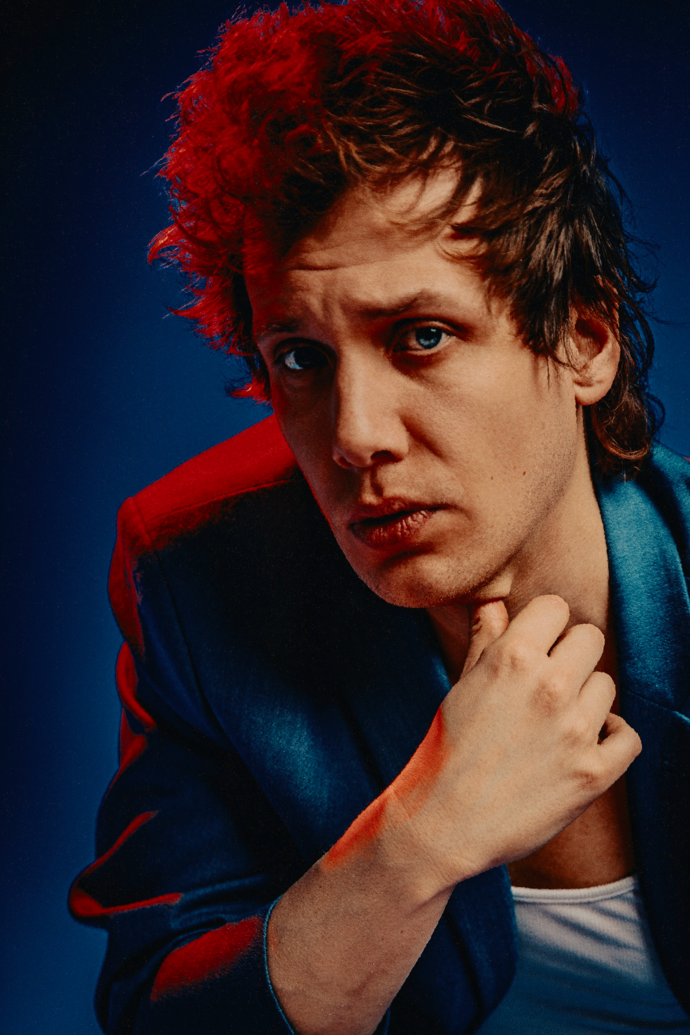 Curly red-haired young man in blue jacket posing intensely against deep blue background.
