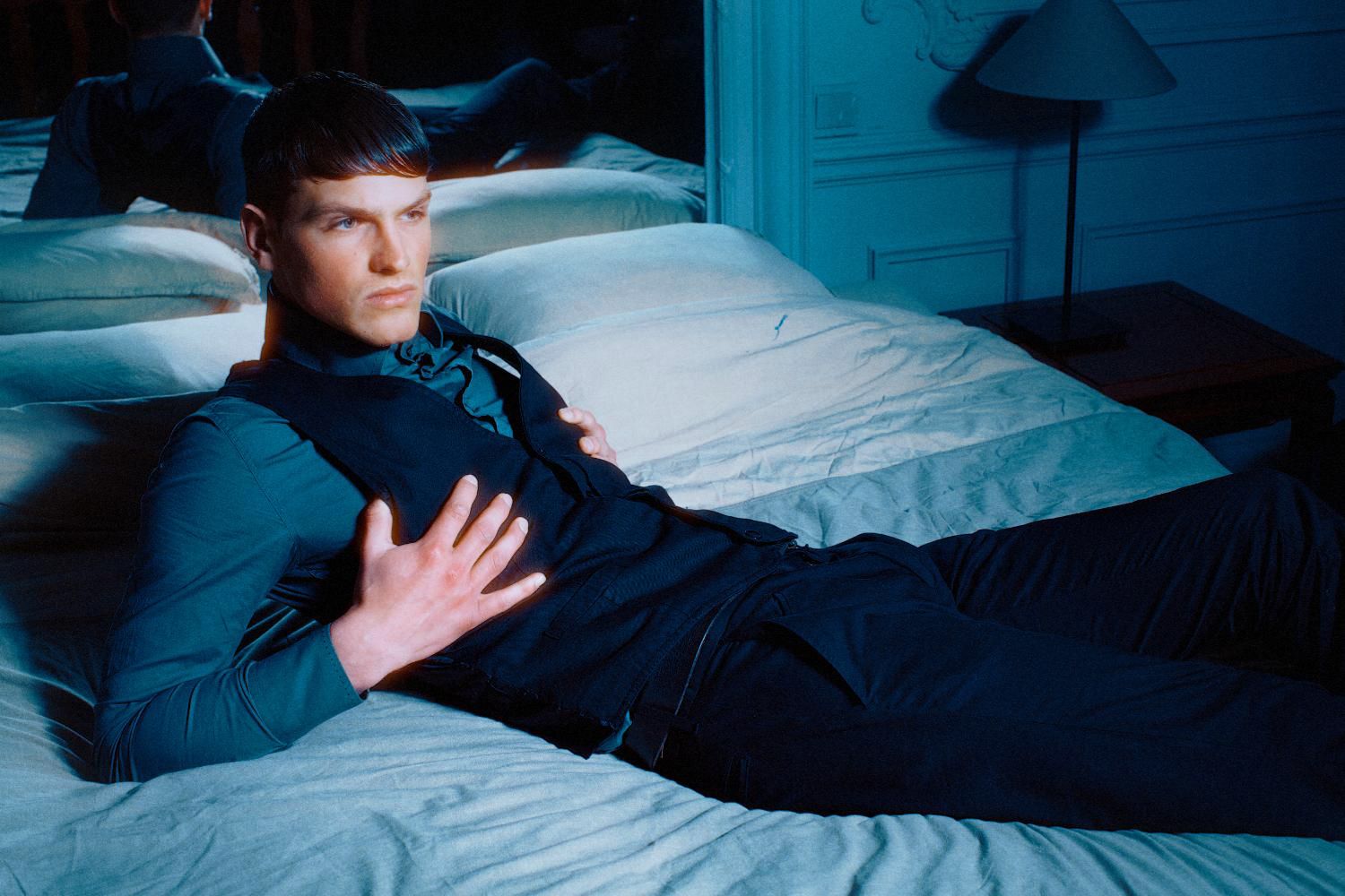 Man in dark clothing reclines on bed in moody blue-lit bedroom setting.