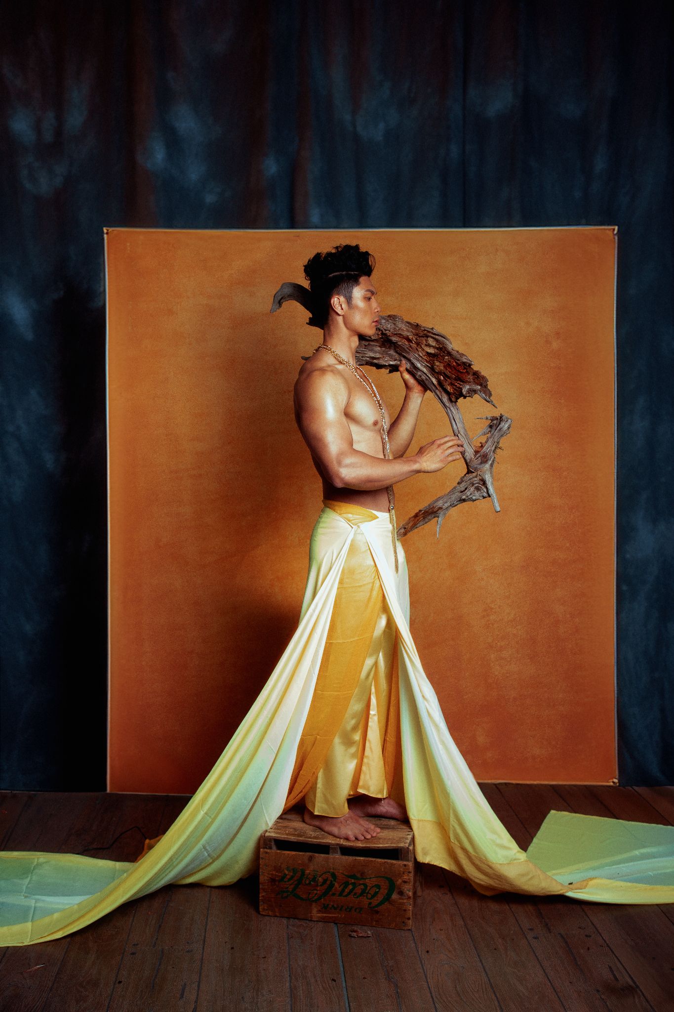 Shirtless Black male model in golden yellow draped skirt holds dried botanicals against orange studio backdrop.
