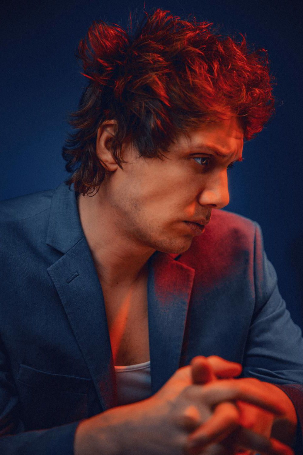 Brooding man with curly red-lit hair wearing blue blazer in dramatic red and blue studio lighting.