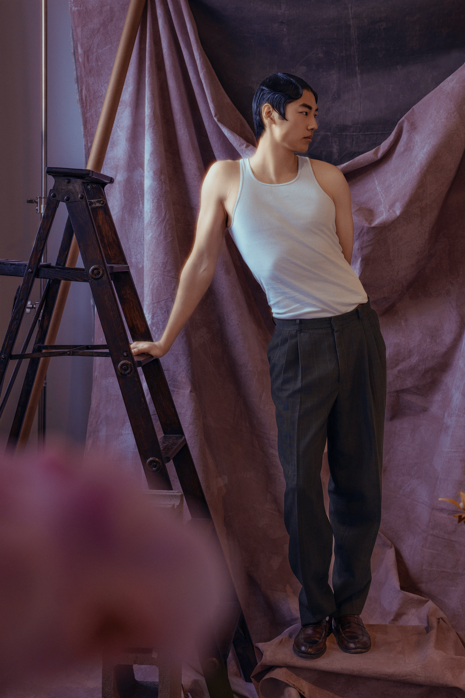 Athletic man in white tank top and grey trousers poses beside wooden ladder with draped fabric backdrop.