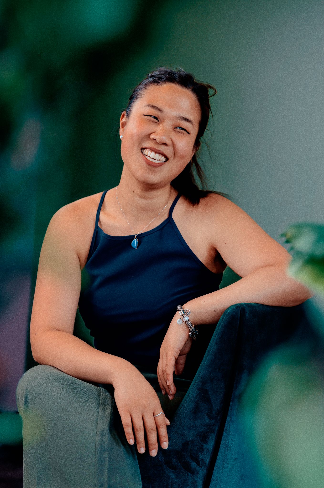 Smiling woman in navy tank top seated casually against a green leafy background.