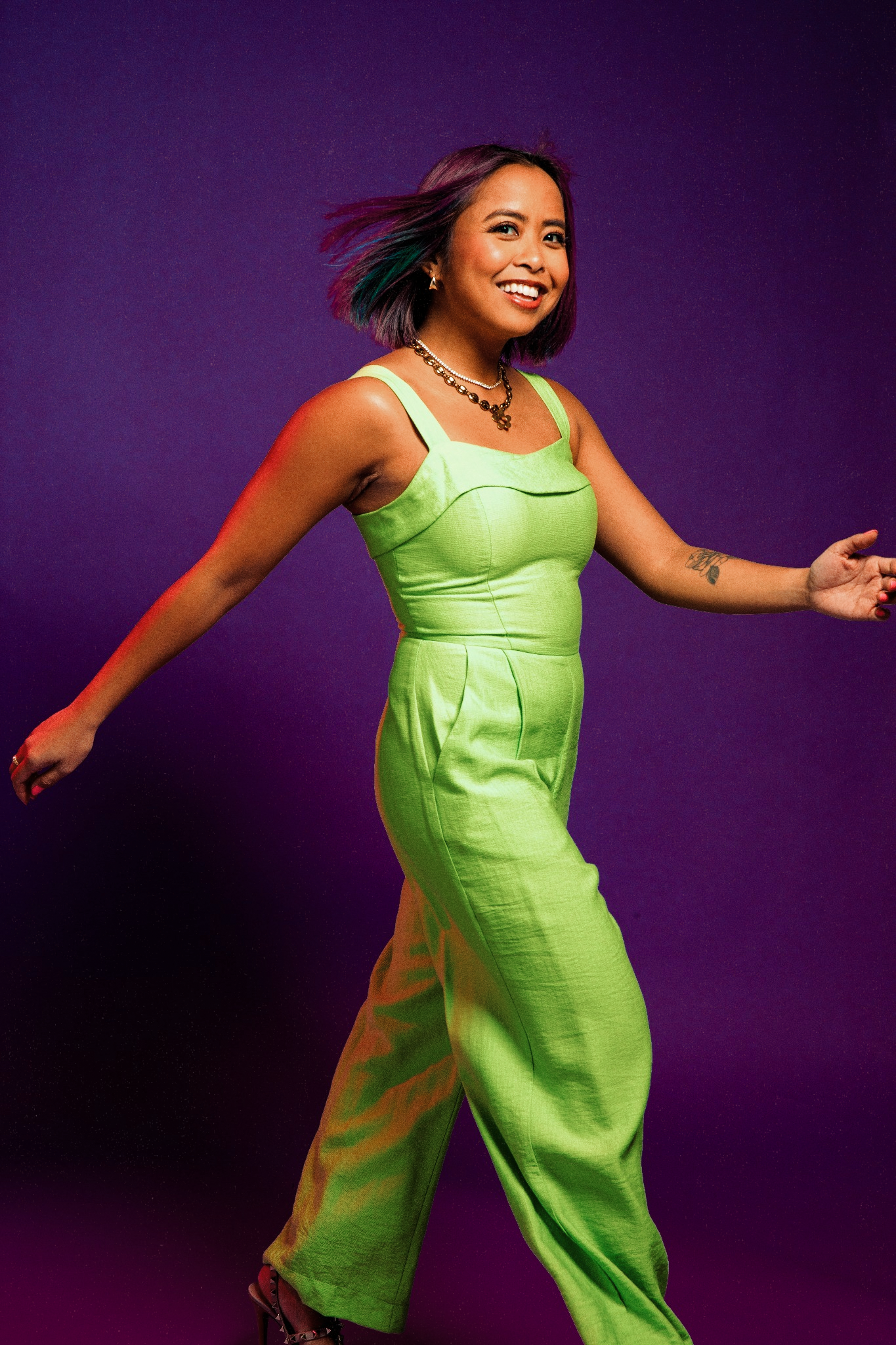 Smiling woman in mint green jumpsuit posing dynamically on a purple background.