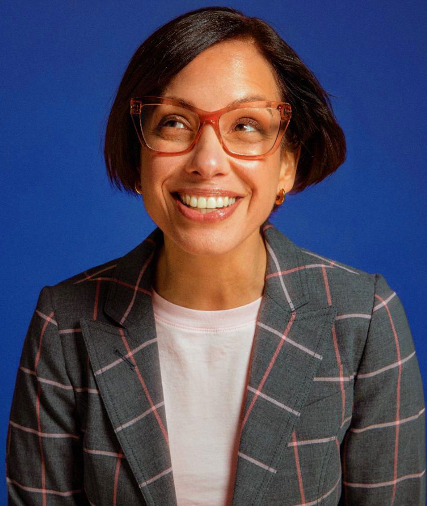 Cheerful woman with short hair wearing red glasses and plaid blazer smiling against blue background.