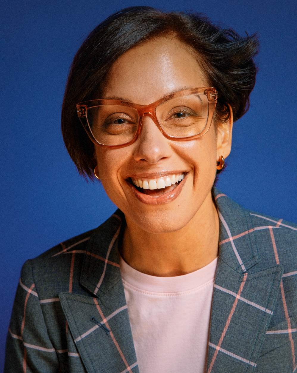 Woman with short hair wearing plaid blazer and orange glasses laughs against bright blue background.