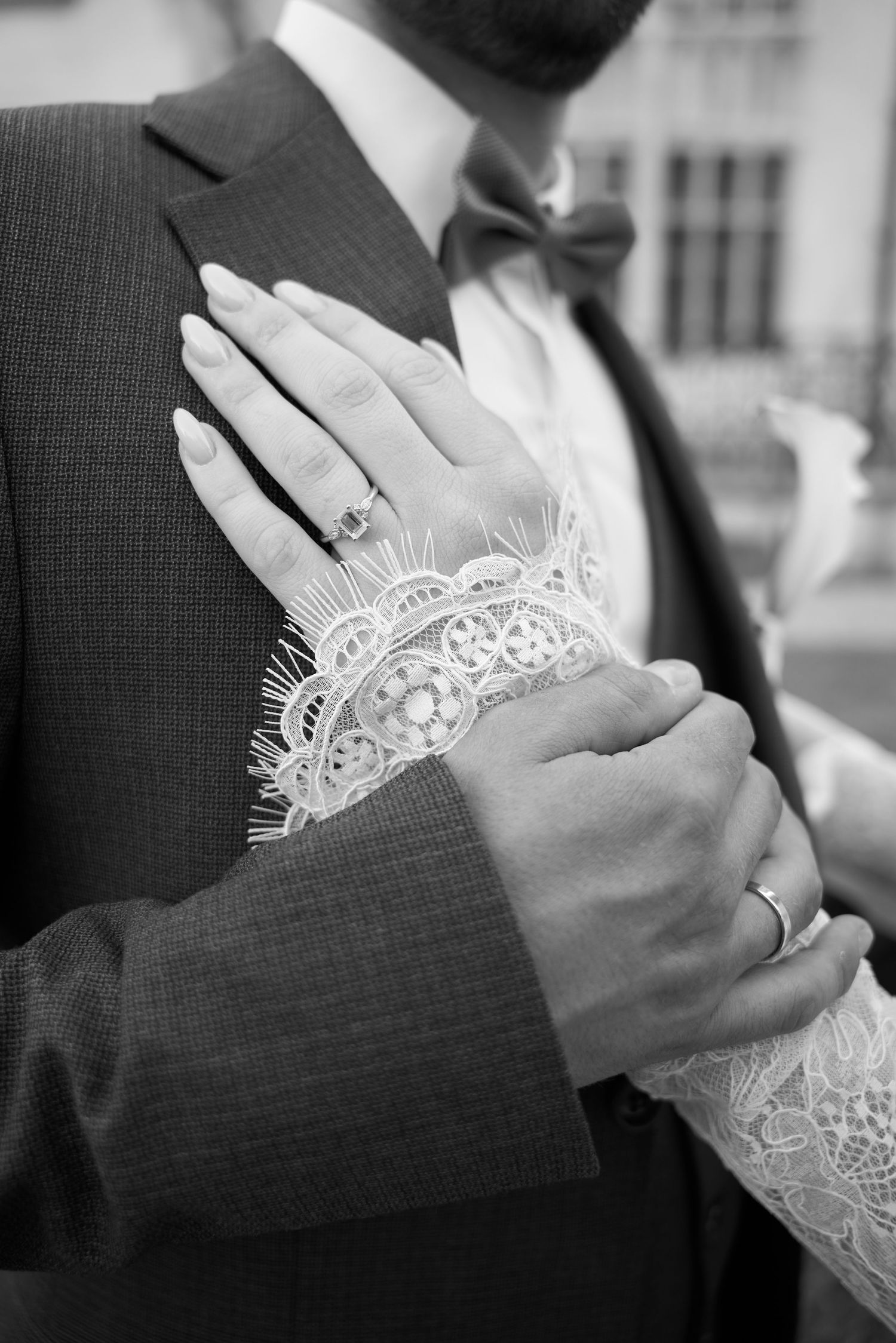 Black and white close-up of bride and groom hands showing wedding rings, lace bridal sleeve detail.