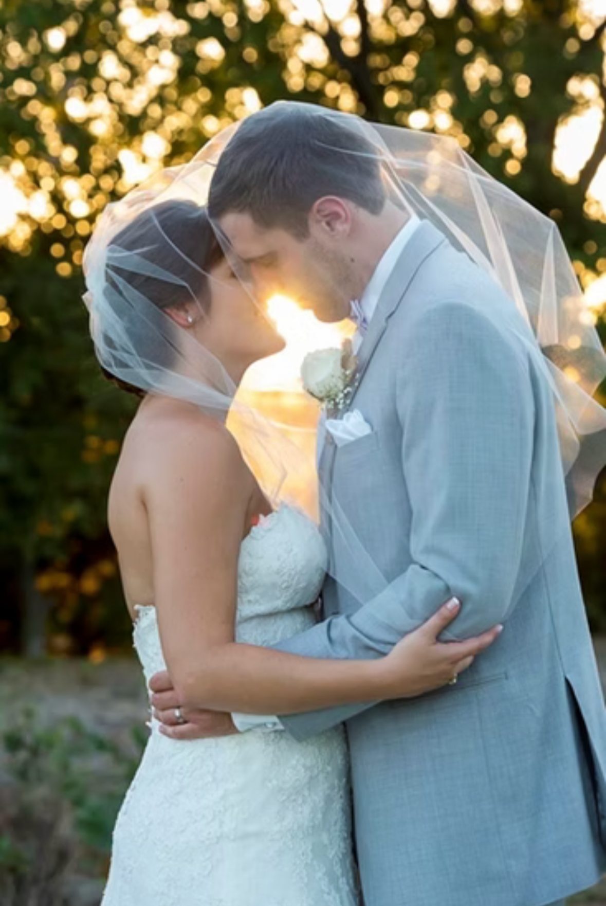 Bride and groom sharing intimate moment under veil with golden sunset backlight in outdoor setting.