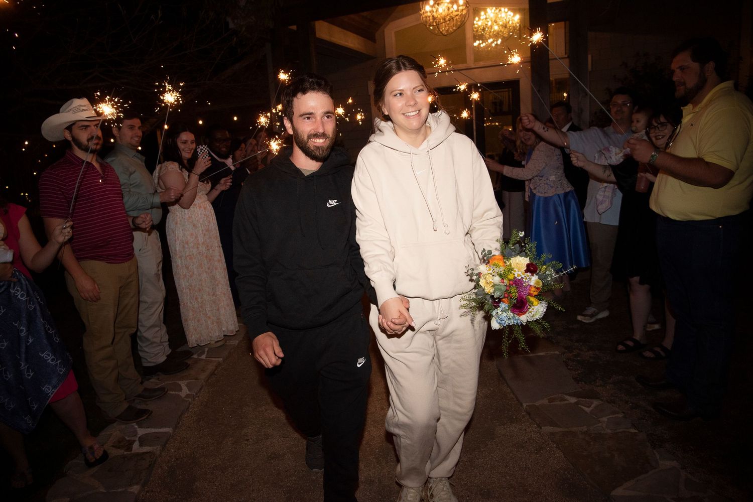 Two grooms walk hand-in-hand through a sparkler exit tunnel at their outdoor wedding reception at night.