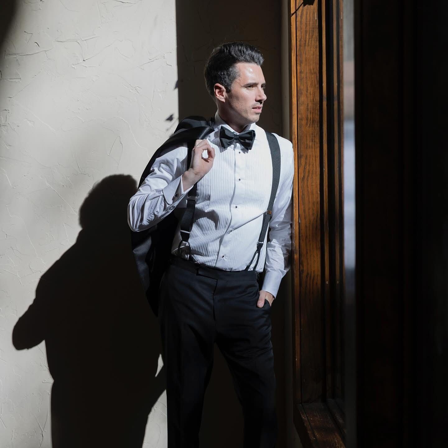 Stylish groom in white shirt, suspenders and bow tie leaning against wooden door in sunlight at the Springs in Red Oak Texas