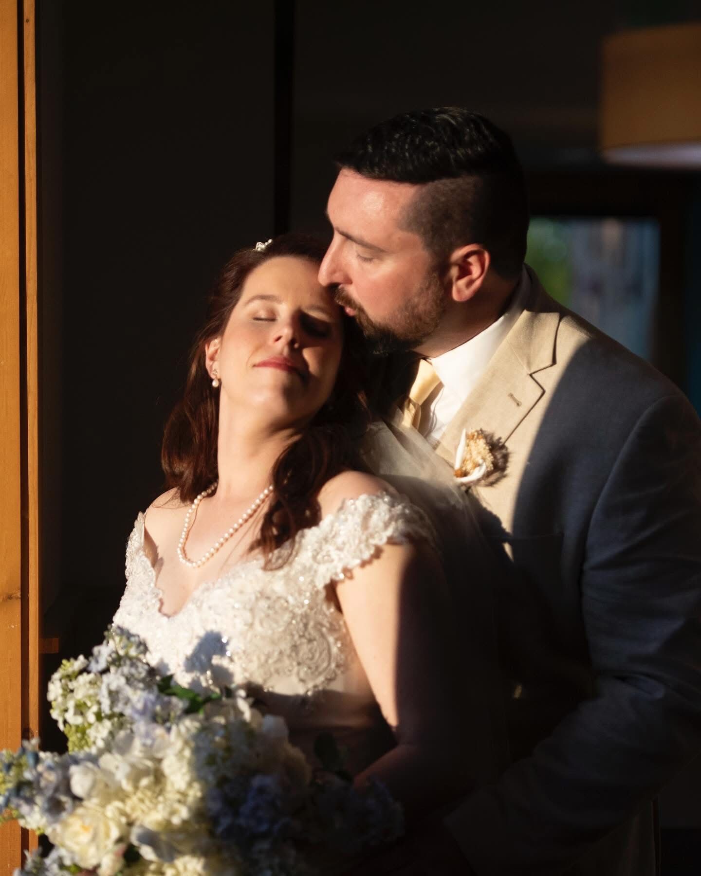Bride and groom share a tender moment as groom kisses bride's temple while she holds blue floral bouquet.