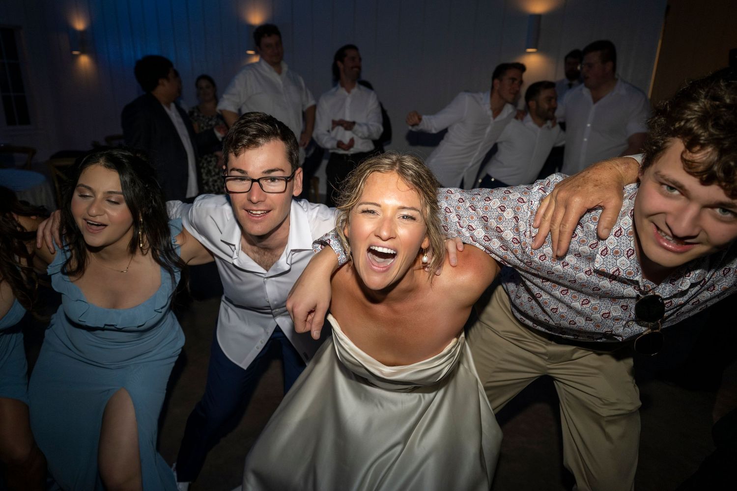 Bride laughs joyfully with wedding guests during energetic dance floor celebration at indoor reception.