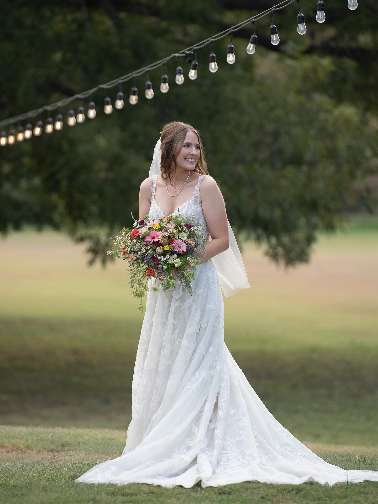 Radiant bride in lace gown holds colorful bouquet while standing under string lights at Stonebridge Venue in Blum Texas
