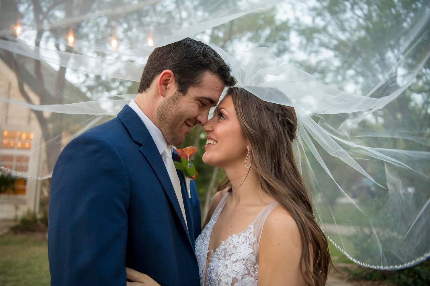 Romantic bride and groom gazing at each other under flowing veil at outdoor wedding reception.