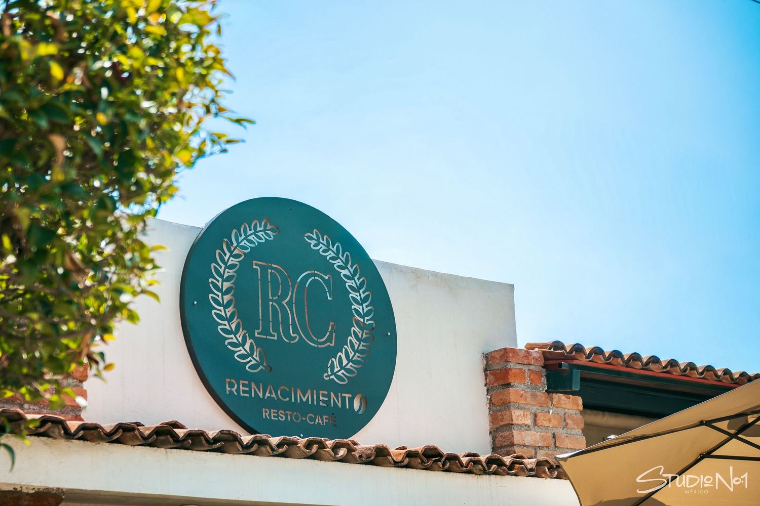 RC Renacimiento Mexican restaurant circular green logo sign mounted on white stucco wall with terracotta roof tiles.