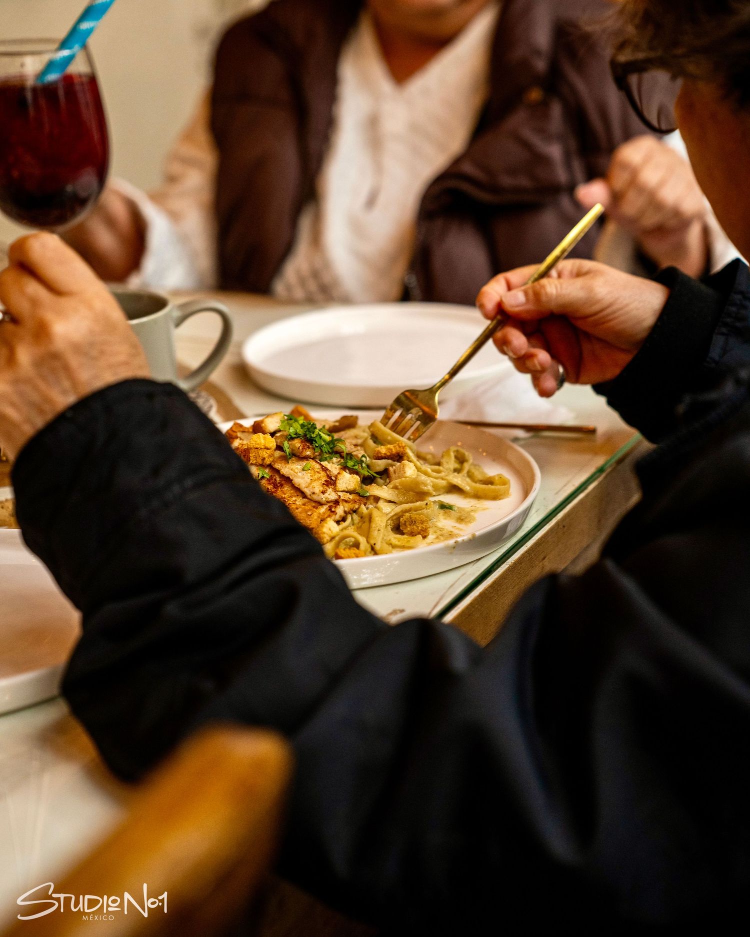 People dining at a restaurant table, sharing pasta dishes with forks, red wine glass visible nearby.