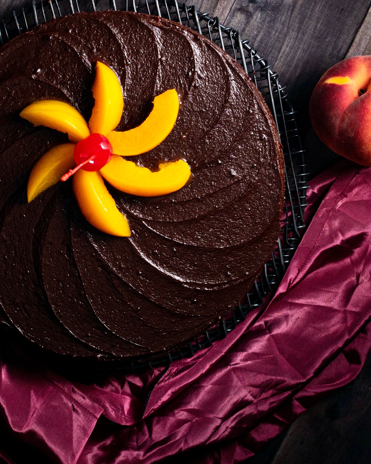 Decadent chocolate cake topped with fresh peach slices and a cherry on a cooling rack.