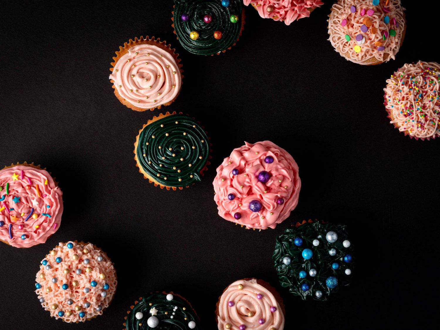 Assorted colorful cupcakes with pink, teal frosting and sprinkles arranged on a dark background.