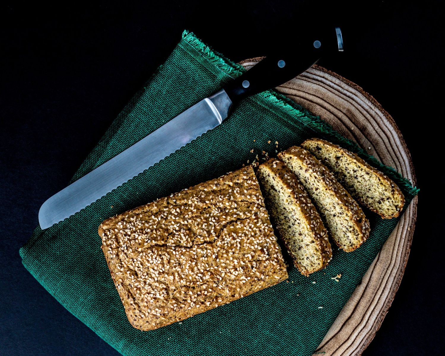 Sliced seeded loaf bread on a green cloth with a bread knife in a wooden basket.
