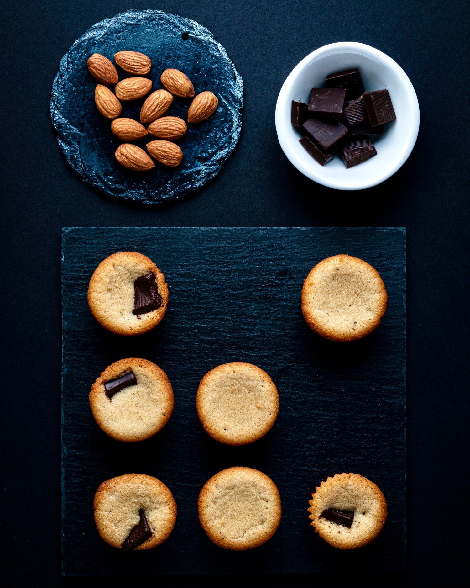 Flat lay of almond chocolate mini tarts with almonds and dark chocolate pieces on a slate board.