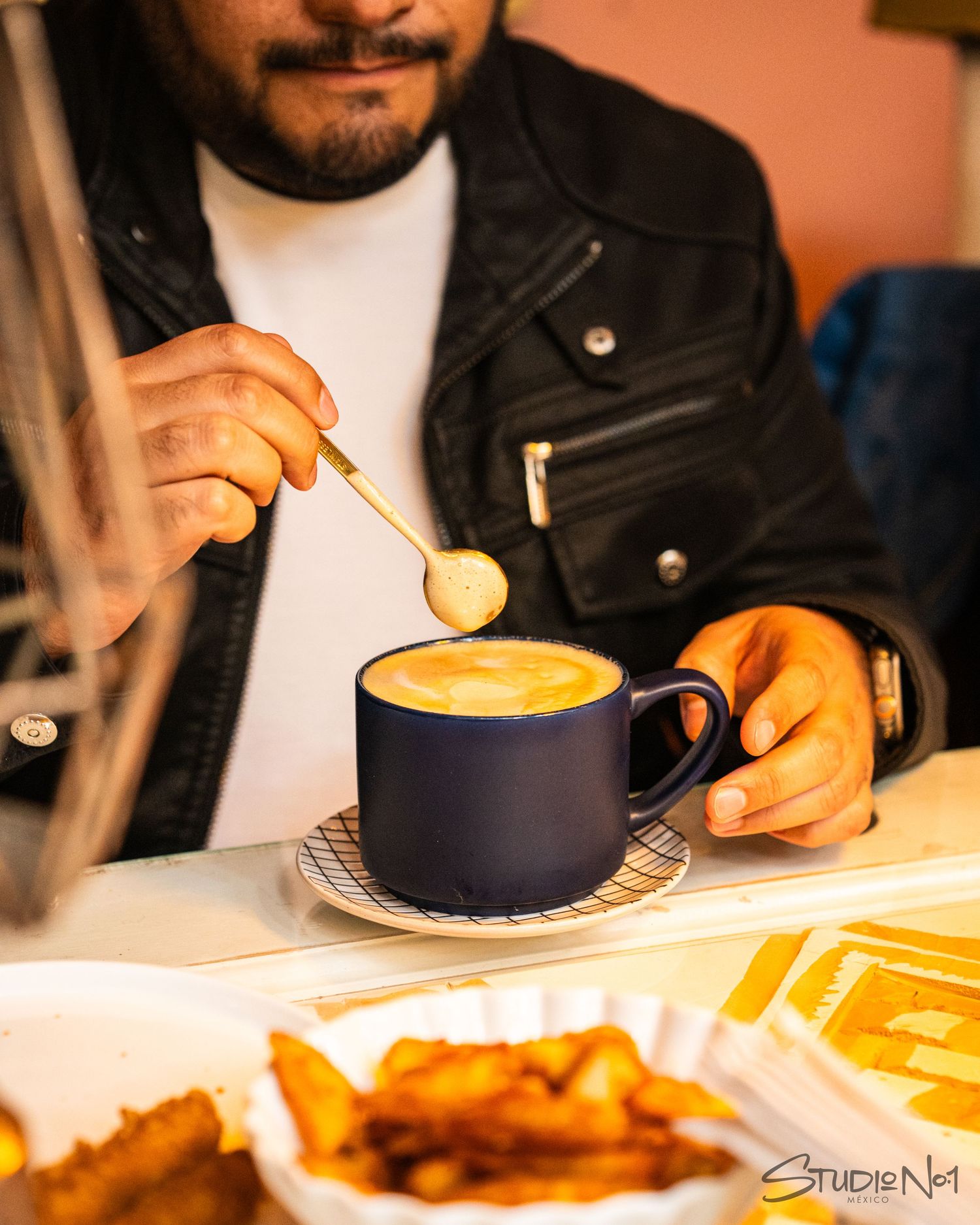 Man in leather jacket stirring frothy coffee in a blue mug at a cafe table with fries nearby.