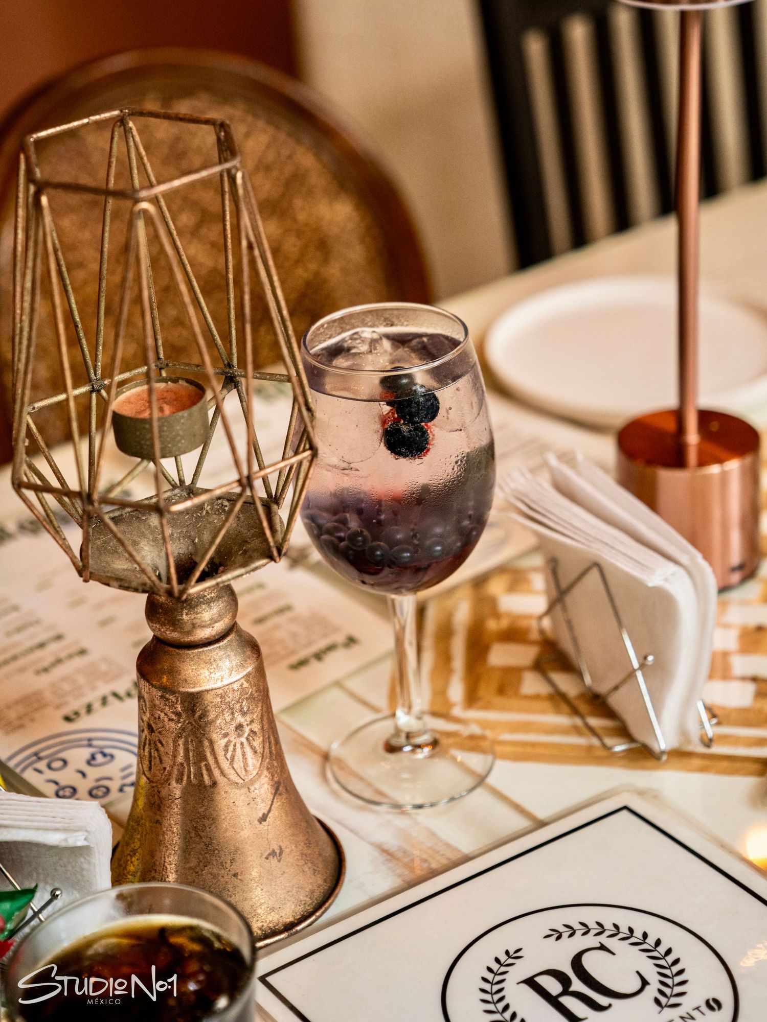 Elegant restaurant table with a geometric brass candle holder, blueberry cocktail in wine glass, and RC branded placemat.