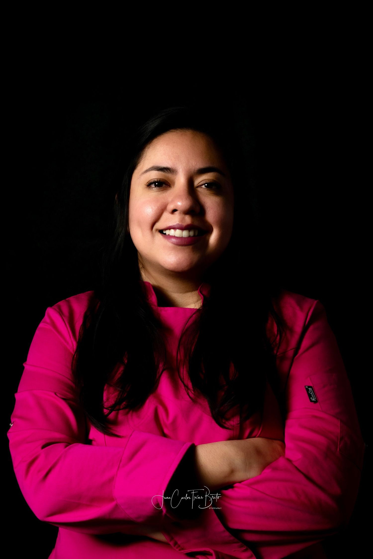 Portrait of a smiling female pastry chef in a pink uniform with arms crossed against a black background.