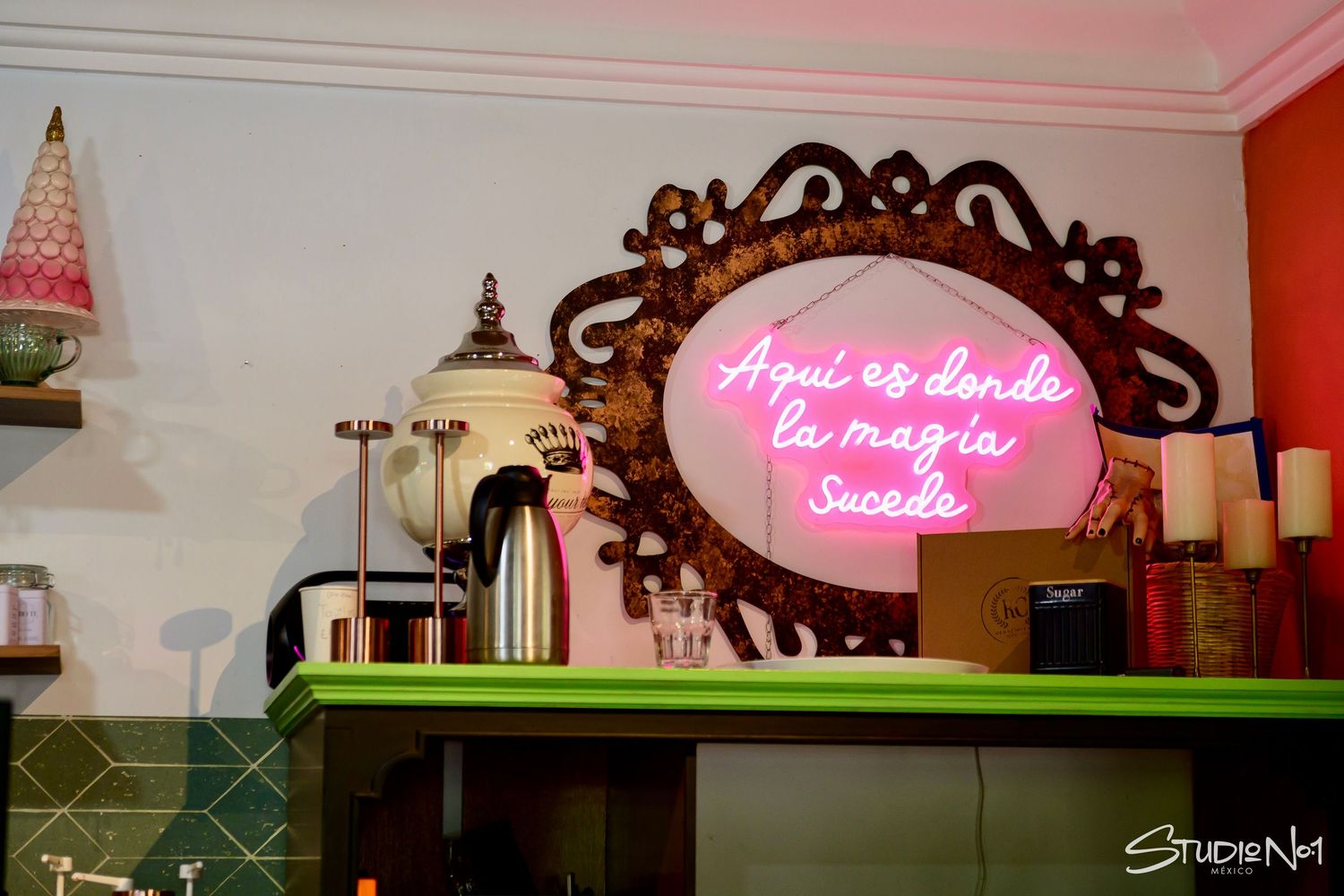 Neon pink sign reading Aqui es donde la magia sucede in ornate mirror above green counter in cozy cafe.