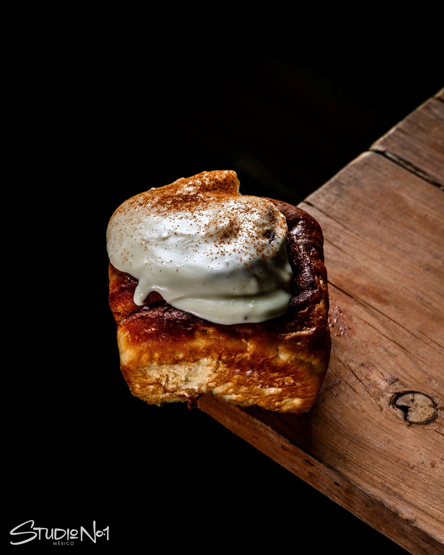 Toasted cinnamon roll topped with whipped cream and cinnamon on a rustic wooden surface against dark background.