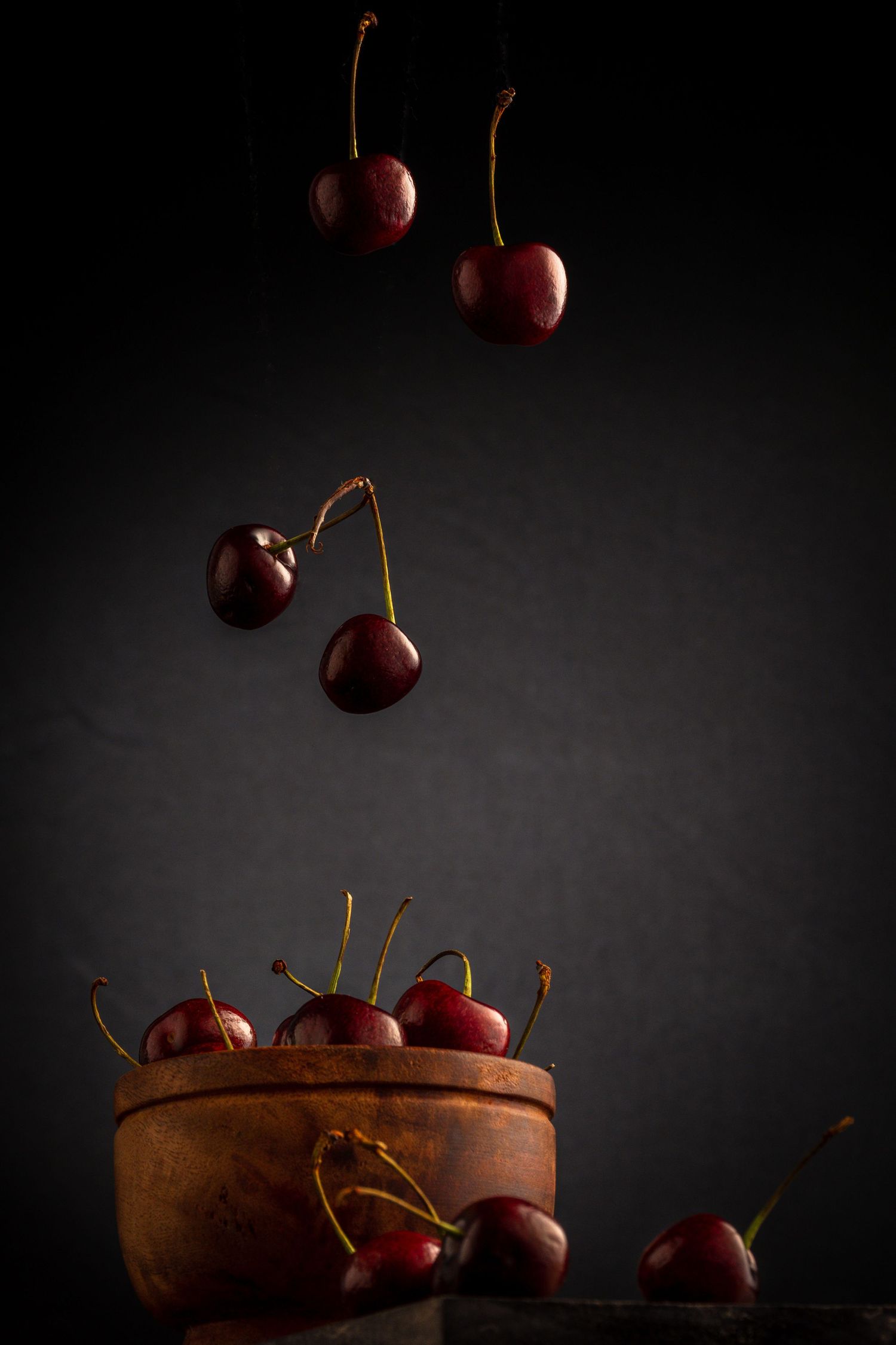 Dark moody photo of fresh red cherries falling into a rustic wooden bowl against a dark background.