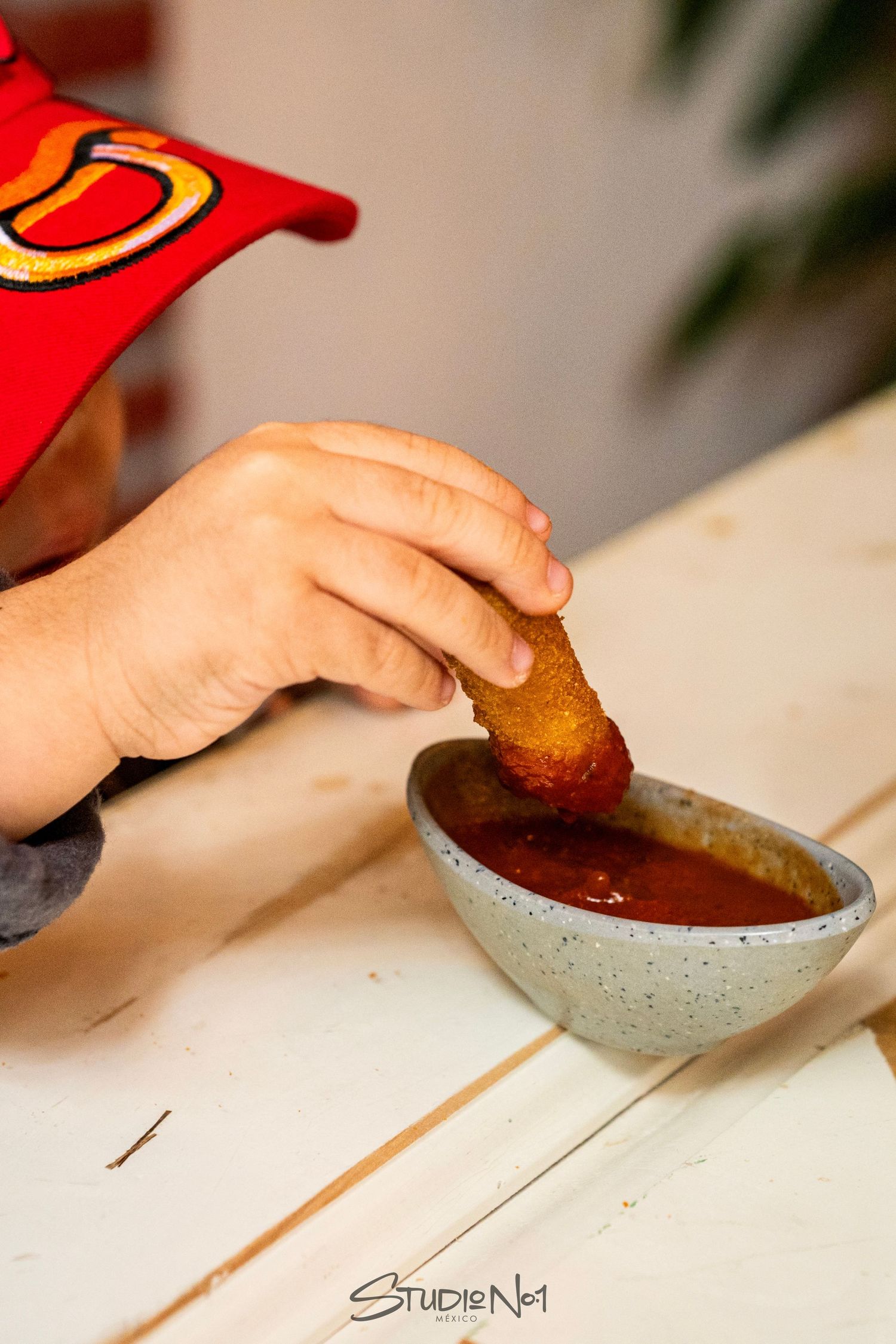 Child dipping a golden fried nugget into a bowl of red tomato sauce on a white wooden table.