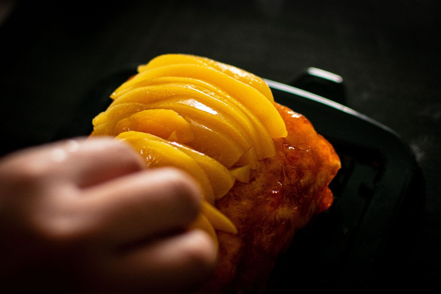 A hand holding a golden mango tart with layered sliced mango on top over a dark plate.