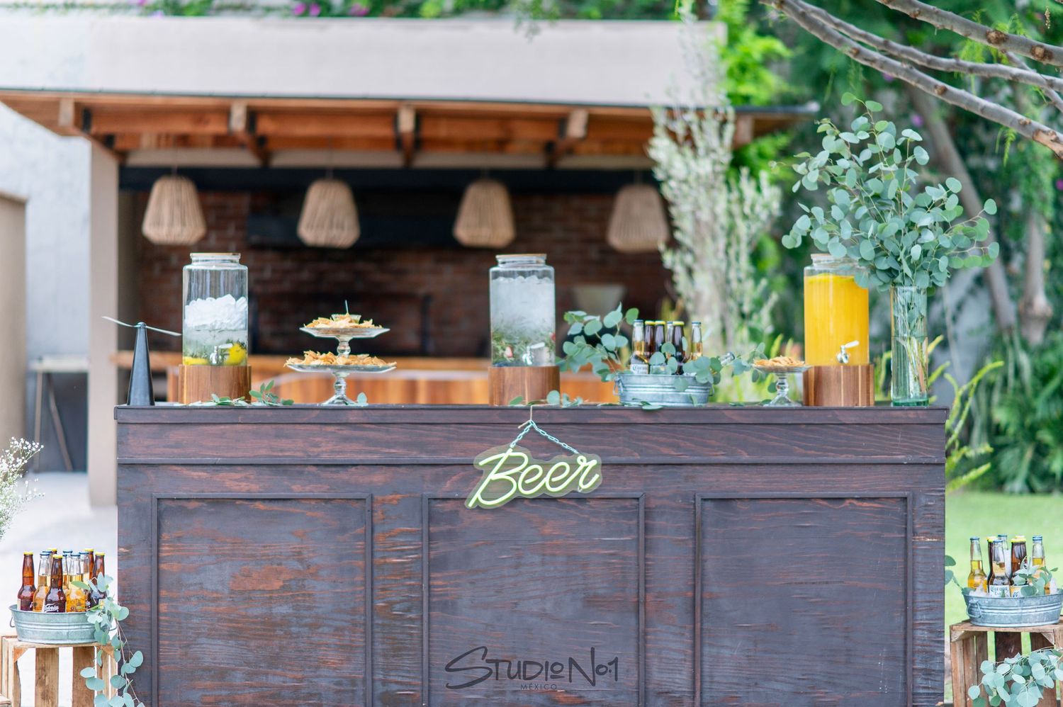 Rustic wooden outdoor bar station decorated with greenery and labeled Beer sign at a wedding.