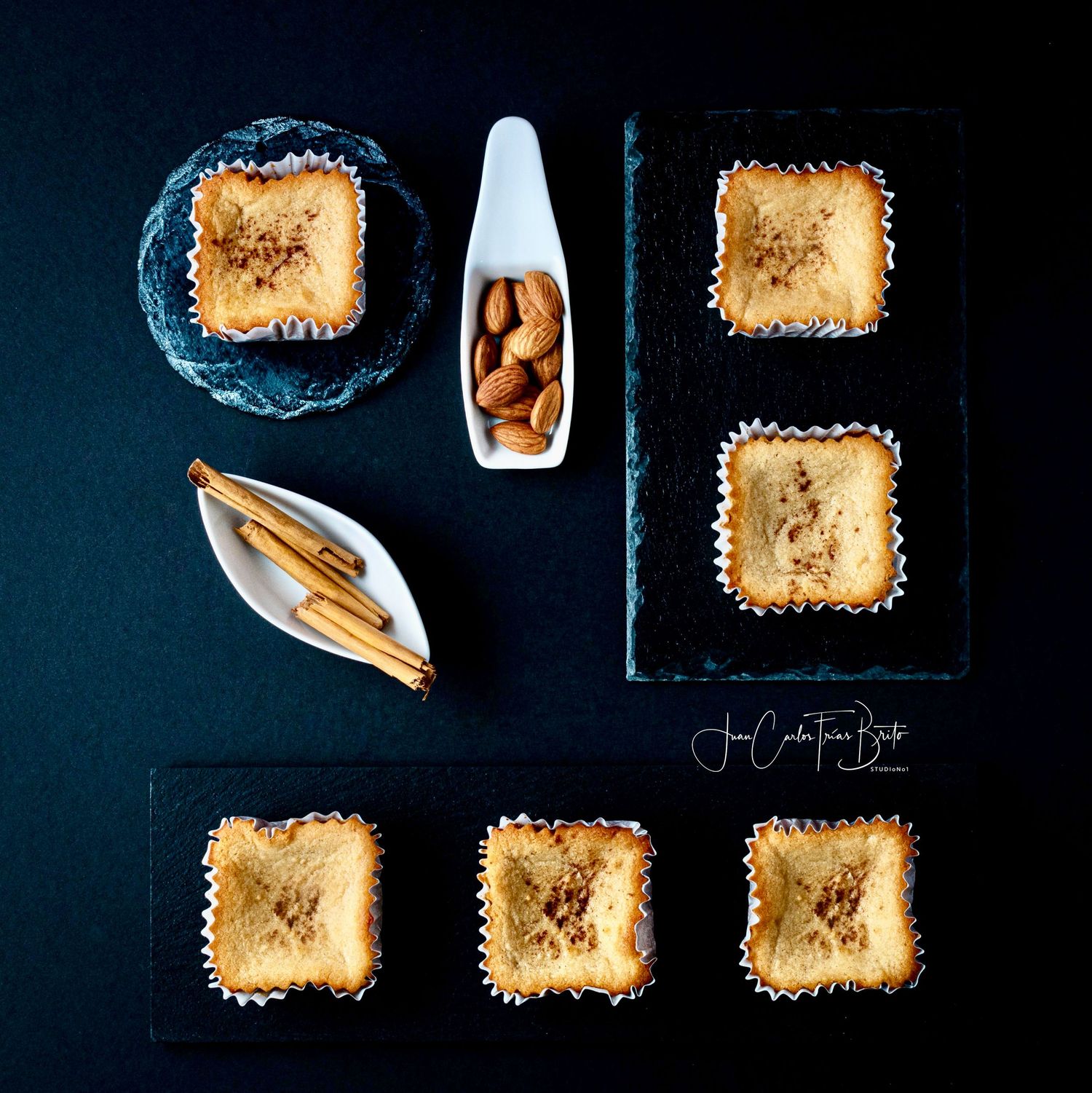 Flat lay of square almond pastries with cinnamon sticks and almonds on dark slate boards.