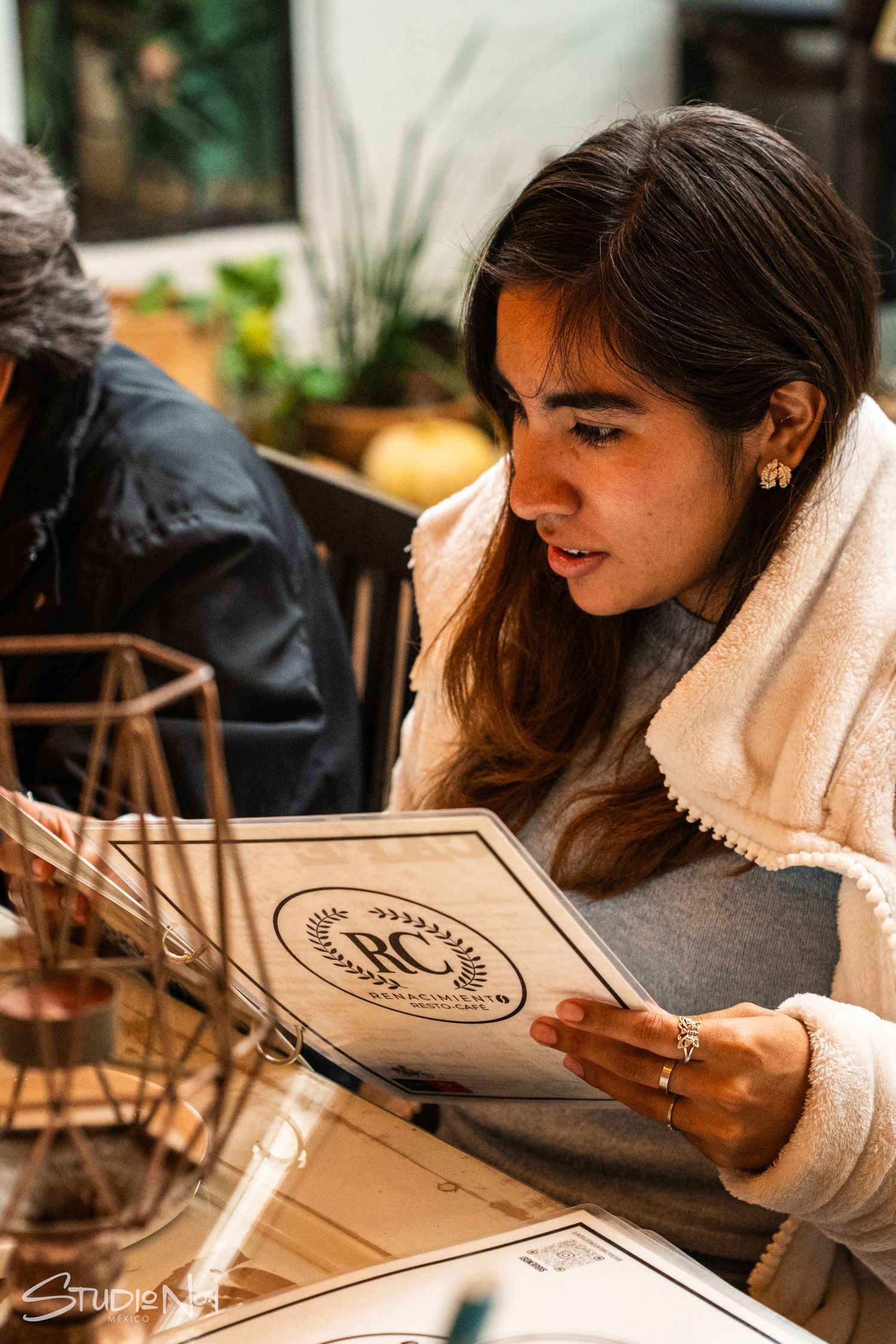Young woman reading a restaurant menu at a cafe table, wearing a cozy white jacket and gold earrings.