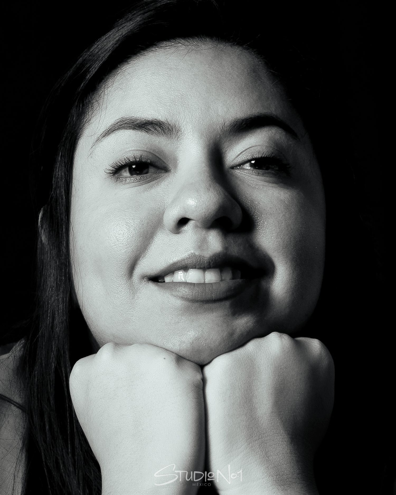Black and white portrait of a smiling young woman resting her chin on her fists against a dark background.