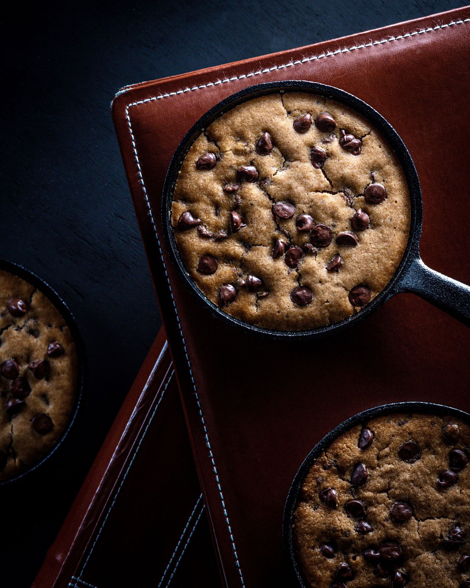 Three cast iron skillets with warm chocolate chip cookies on a dark background.