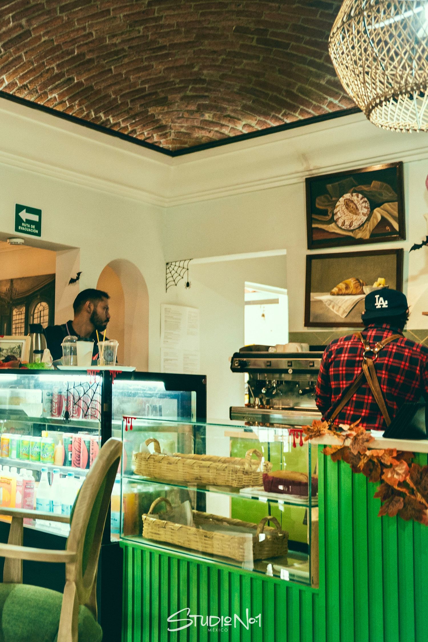 Interior of a cozy cafe with green display counter, brick ceiling, artwork on walls, and two baristas preparing drinks.