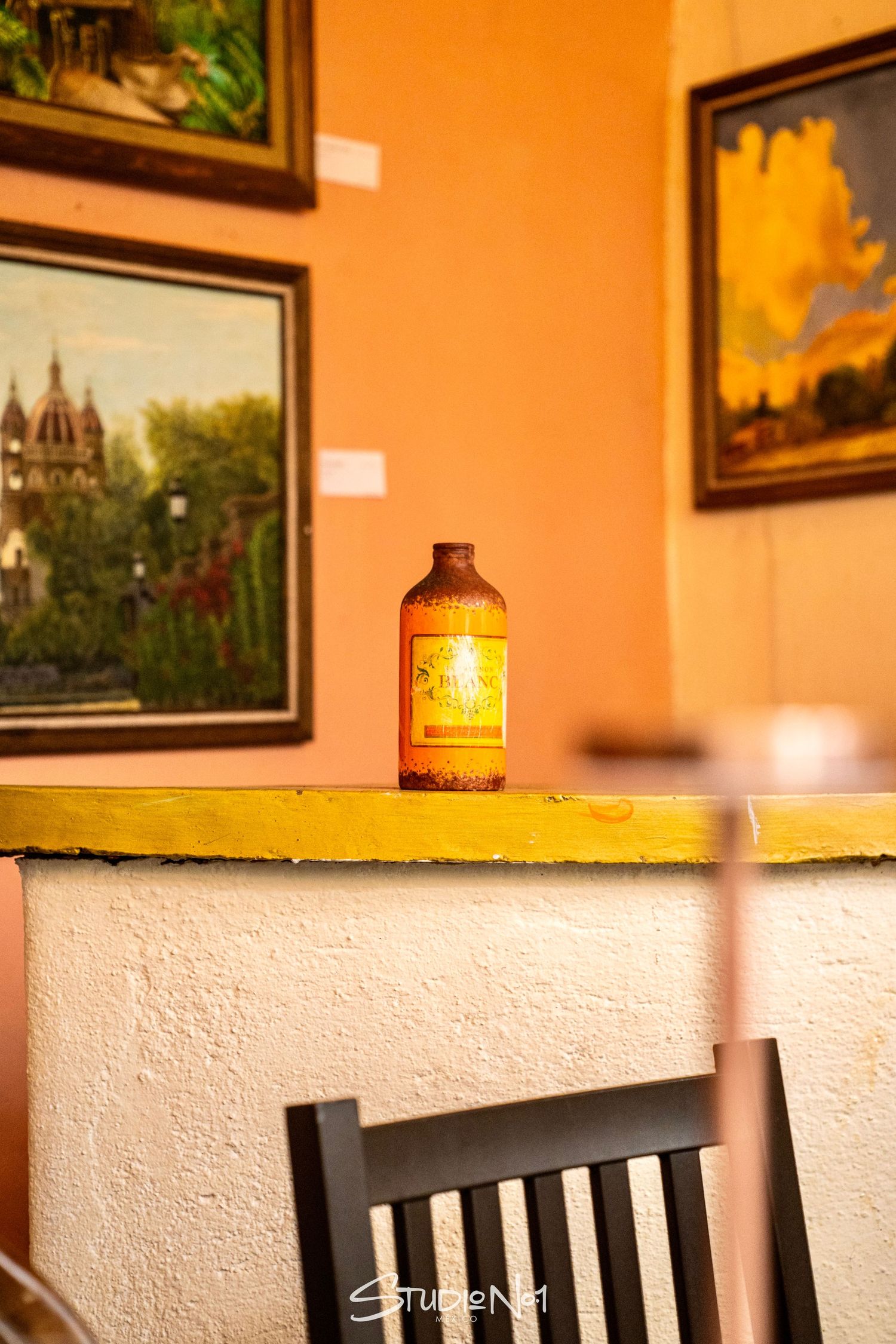 Vintage bottle on yellow shelf with orange walls and framed landscape paintings in a cozy cafe setting.