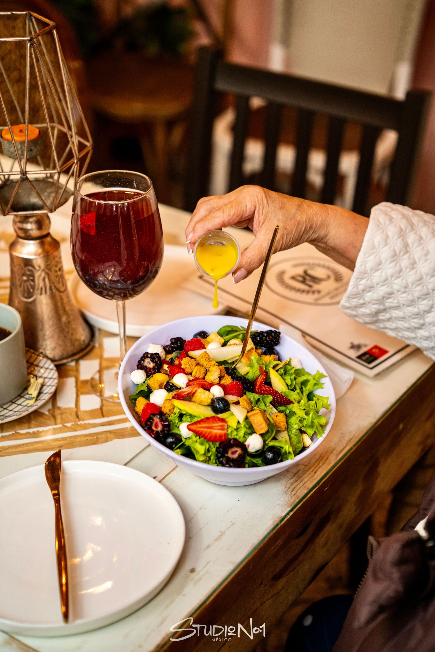 Person drizzling dressing over a colorful fresh salad with berries and mozzarella, beside a glass of red wine.