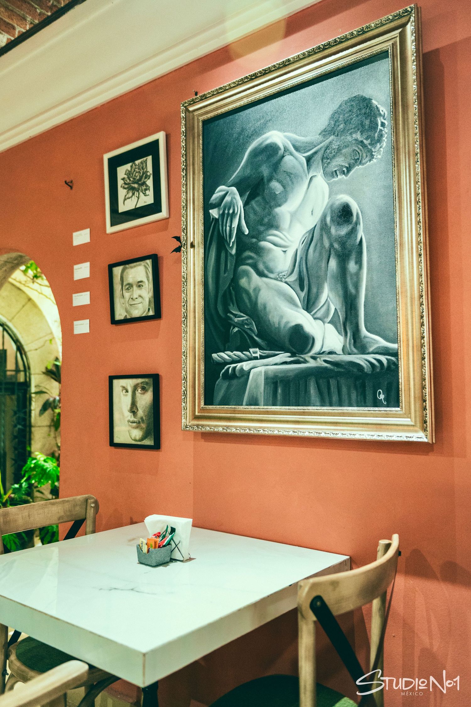 Cozy cafe interior with salmon walls, large black and white figurative painting, and smaller portrait artworks above a white table.