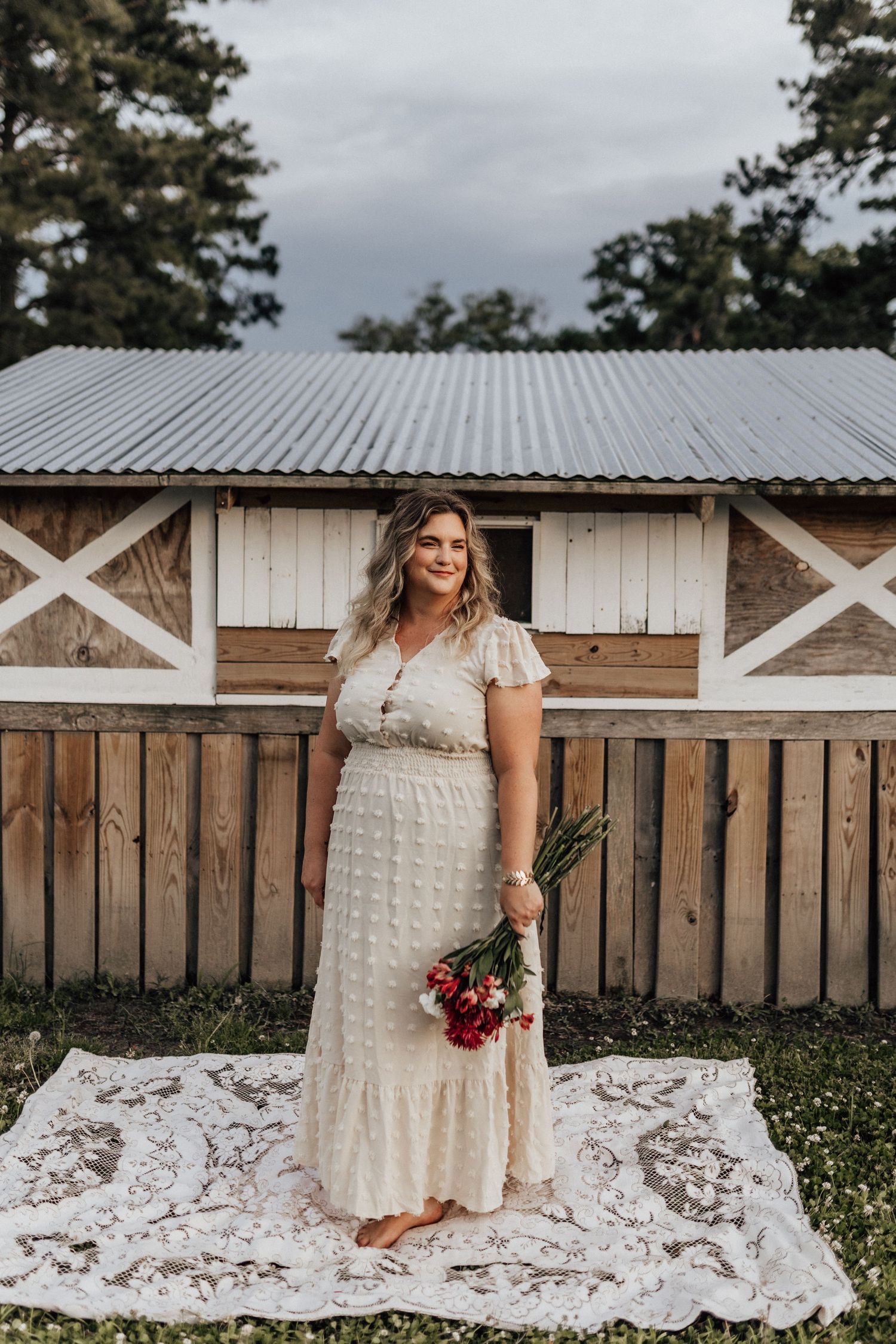 Headshot of the brand photographer in a cream lace maxi dress stands on a white lace blanket holding red flowers in front of a rustic barn.