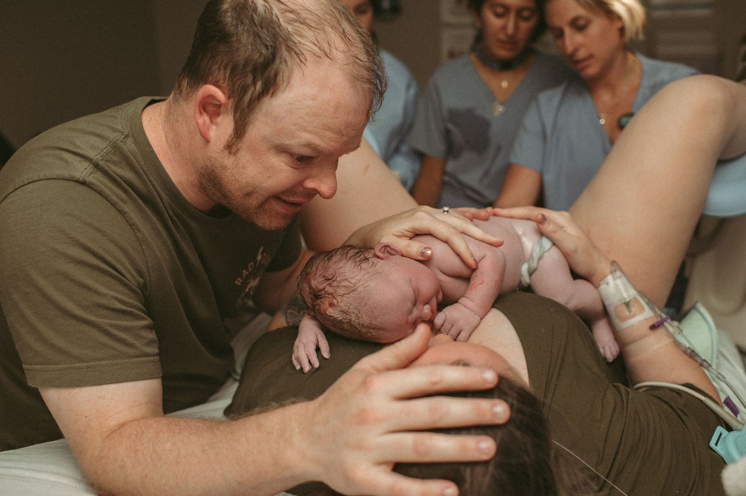 Father tenderly holds newborn baby on mother's chest in hospital delivery room after birth in Norfolk, Virginia.