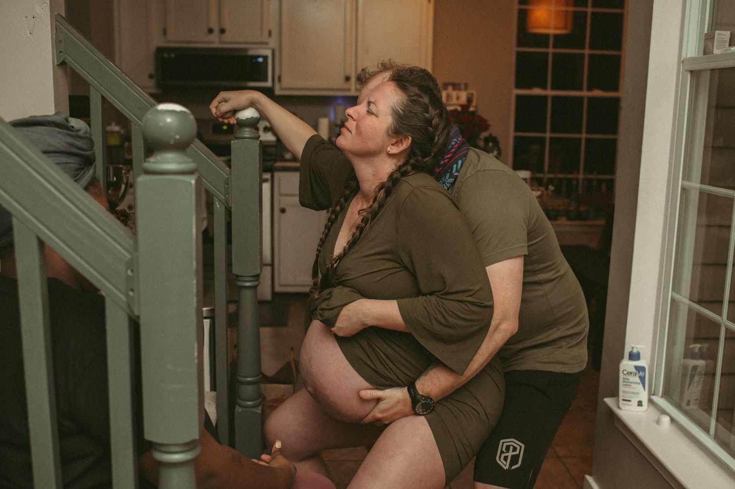 Pregnant woman kneeling holding her belly while laboring against staircase railing in her home located in Chesapeake, VA.