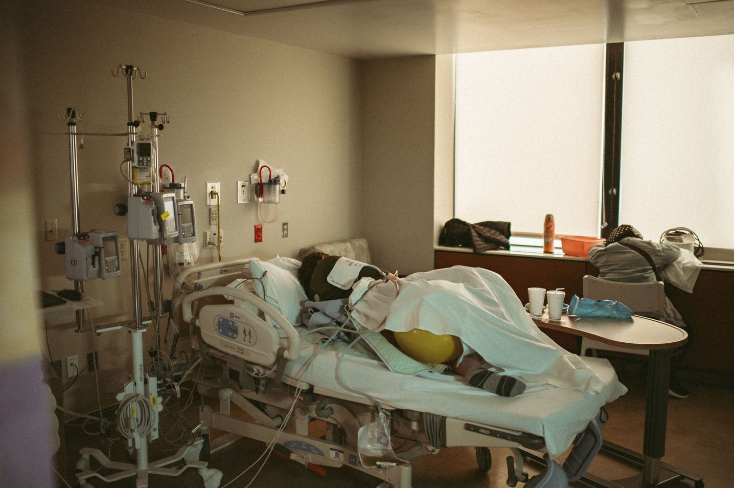 Expecting mom resting in hospital bed surrounded by medical equipment in a dimly lit private room with large window in Norfolk, Virginia.
