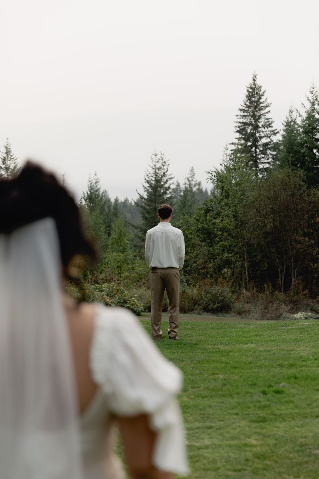 Bride in white dress approaches groom standing alone on green lawn facing tall evergreen trees.